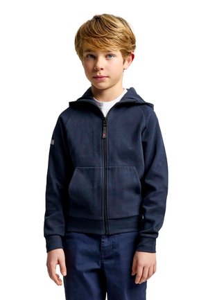Un niño está de pie delante de un fondo blanco, vestido con una sudadera con capucha azul marino y cremallera y unos pantalones azul oscuro.