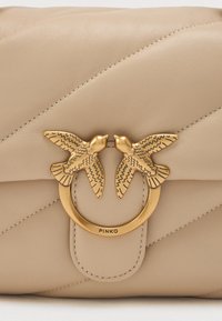 Beige bőr kézitáska steppelt textúrával, amelyen aranyszínű köralakú csatt található, két madármotívummal és logó részlettel az elején.