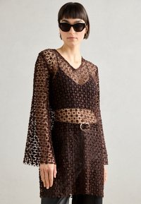 Brown lace tunic with long sleeves, featuring a floral pattern. A semi-transparent overlay, black underlayer, and a belt accent at the waist.