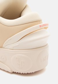 Sneaker beige con una parte superiore testurizzata, suola bianca spessa con loghi in rilievo e un accento rosa sulla linguetta del tallone. Dettagli di design unici inclusi.