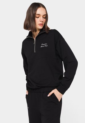 Sweatshirt noir zippé en coton doux, avec un col monté, des poignets côtelés et un texte brodé sur la poitrine. Texture lisse.