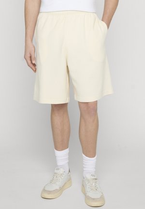 Mężczyzna noszący teksturowane, kremowe szorty, białe skarpetki do kostek i białe trampki na jednolitym tle, z jedną ręką w kieszeni.