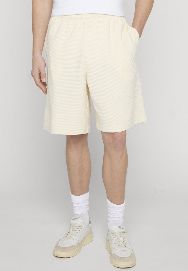 Férfi, aki törtfehér, texturált rövidnadrágot, fehér bokazoknit és fehér tornacipőt visel, egyszerű háttér előtt, egyik kezét a zsebében tartva.