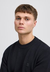 Fiatal férfi, rövid barna hajjal és kék szemmel, fekete bordás pulóverben és kis fülbevalóval egy sima háttér előtt.