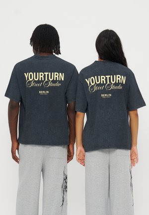 Zwei Personen stehen nebeneinander mit dem Rücken zur Kamera, tragen dunkle "YOURTURN Street Studio Berlin" T-Shirts und hellgraue Hosen mit schwarzen Mustern.
