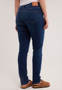 Persoană purtând blugi slim fit albastru închis și sandale maro slip-on, stând cu spatele la un fundal neutru.