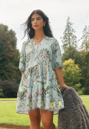 Frau in einem hellblauen Blumenkleid, die einen grauen, strukturierten Mantel hält, steht im Freien vor Bäumen und Gras.