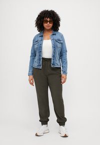 Chaqueta de mezclilla con cierres de botones sobre una camiseta blanca, combinada con joggers olivo y zapatillas blancas. Presenta un corte holgado, puños elásticos y bolsillos laterales.