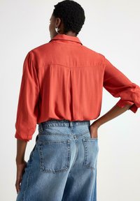 Rustrød, let button-up skjorte med løs pasform og optrukne ærmer, parret med højtaljede, falmede blå denimbukser.