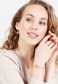 Gouden metalen sieradenset met een hangerketting en oorringen met een logo ontwerp. Het model draagt een lichtbeige top en toont de accessoires.