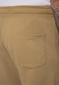 Beige Baumwoll-Sweatpants mit geripptem Bund und einer Gesäßtasche mit einem dreieckigen Akzent. Glatte Textur und schlichtes Design.