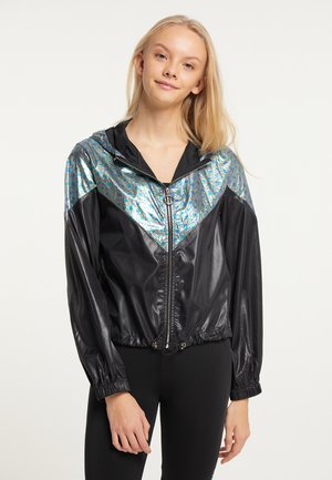 Junge Frau mit langen blonden Haaren, die eine schwarze Jacke mit einem glänzenden silber-blauen gemusterten oberen Abschnitt und schwarzen Leggings trägt.