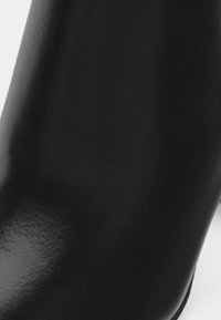 Gros plan d'une botte en cuir noir lisse avec une légère texture et un doux reflet de lumière sur la surface courbée.