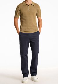 Beige gestricktes Poloshirt mit Kragen, kombiniert mit marineblauen Hosen und weißen Sneakers, die eine strukturierte Oberfläche und einen klassischen Schnitt zeigen.
