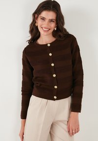 Gilet rayé marron avec un motif texturé, comportant de gros boutons dorés sur le devant, associé à un pantalon clair.