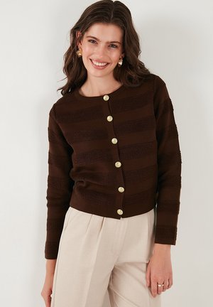 Gilet rayé marron avec un motif texturé, comportant de gros boutons dorés sur le devant, associé à un pantalon clair.