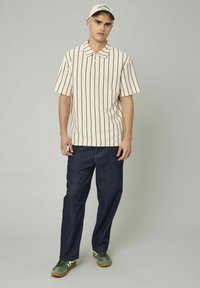 Polo à manches courtes rayé en crème et marine, associé à un jean bleu foncé. Le modèle porte une casquette verte et des baskets vert olive.