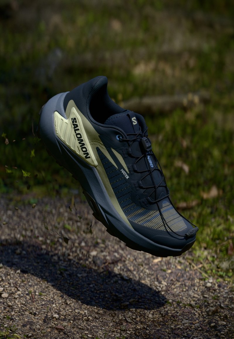 Chaussure de trail Salomon bleu foncé et beige suspendue au-dessus d’un chemin de gravier avec de la verdure en arrière-plan.