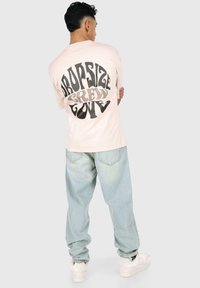 Blassrosa übergroßes T-Shirt mit großen schwarzen und beigen Schriftzügen auf dem Rücken. Kombiniert mit hellblauen weiten Jeans und weißen Sneakers.
