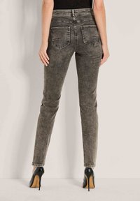 Vrouw die staat, gekleed in vervaagde zwarte skinny jeans met achterzakken en zwarte hoge hakken, tegen een eenvoudige lichte muur als achtergrond.