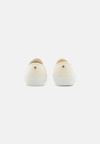 Beige lerretsko med hvite gummisåler, med minimalistisk design og en liten merkeakson bakerst ved hælen.