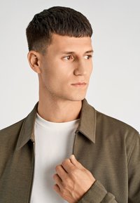 Oliwkowa kurtka zapinana na zamek z kołnierzem, wykonana z teksturowanego materiału. Nosi ją na sobie biała koszulka. Zbliżenie na górną część tułowia i dłoń modela.