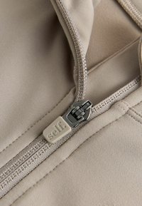 Light beige fabric with a smooth texture, featuring a silver zipper and a square pull tab labeled "self." Stitching is visible along the seam.