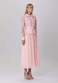 Femme portant une veste en dentelle rose pâle avec des boutons en perles et une ceinture, associée à une jupe midi plissée et des sandales à talons hauts blanches.