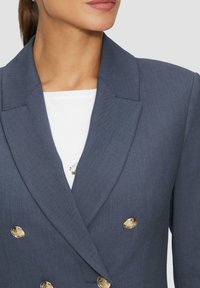 Blazer bleu marine à double boutonnage avec de grands boutons dorés, revers structurés et tissu texturé. Chemise blanche en dessous.