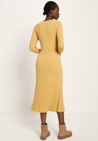 Frau, die seitlich steht und ein senfgelb gestreiftes langärmeliges Midikleid sowie beigefarbene Stiefeletten trägt, vor einem einfarbigen Hintergrund.