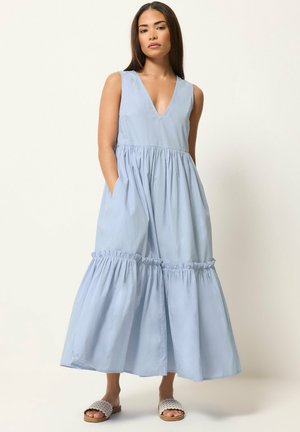 Kobieta stoi, ubrana w jasno niebieską, bezrękawnikową suknię maxi z głębokim dekoltem w kształcie litery V i falbaną na dole, w połączeniu z białymi sandałami wsuwanymi.