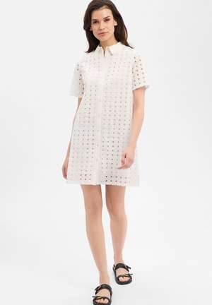 Frau trägt ein weißes, kurzärmeliges Kleid mit Lochmuster und schwarze Sandalen, steht vor einem schlichten, hellen Hintergrund.