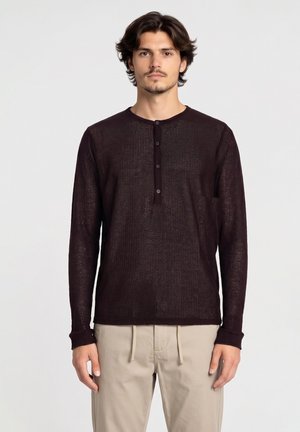 Jonge man met halflang haar draagt een donkerbruin, geknoopt lang mouwen overhemd en beige broek, staand tegen een effen lichte achtergrond.