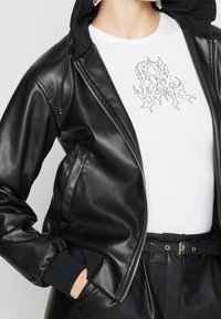 Giacca bomber nera in pelle con chiusura a zip, polsini elasticizzati e cappuccio grigio. Indossata sopra una maglietta bianca con un design grafico nero unico.