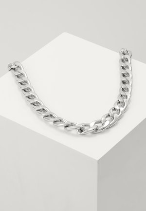 Sølv kæde med store led vist på en hvid geometrisk piedestal mod en ensfarvet grå baggrund.