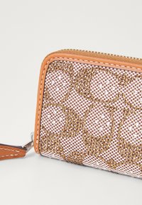 Portefeuille texturé présentant un motif géométrique de perles dorées et blanches sur un fond tissé marron et blanc. Bordures en cuir orange et éléments de fermeture éclair.