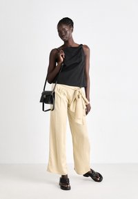 Czarna bluzka bez rękawów, beżowe szerokie spodnie z wiązaniem w talii oraz czarna torebka. Modelka nosi płaskie czarne sandały.