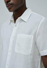 Camisa de lino blanca de manga corta con un acabado texturizado, cierre de botones al frente y un solo bolsillo en el pecho. Presenta un collar clásico y costuras sutiles.