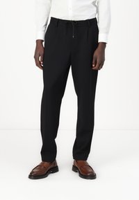 Black tailored trousers with an elastic waistband and drawstring, featuring a straight leg design. Paired with brown leather shoes.