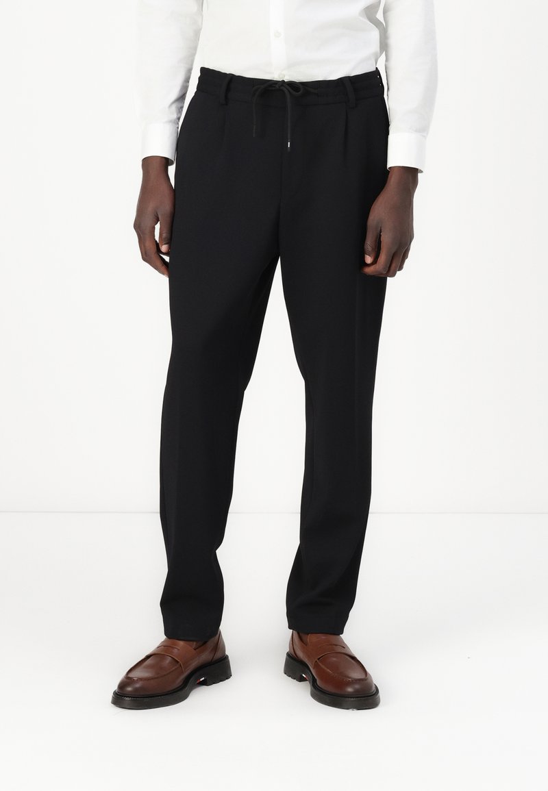 Black tailored trousers with an elastic waistband and drawstring, featuring a straight leg design. Paired with brown leather shoes.