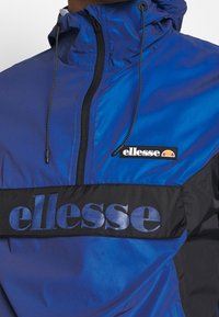 Blue and black windbreaker jacket featuring a hood, zip front, and "ellesse" branding across the chest in bold letters. Satin material finish.