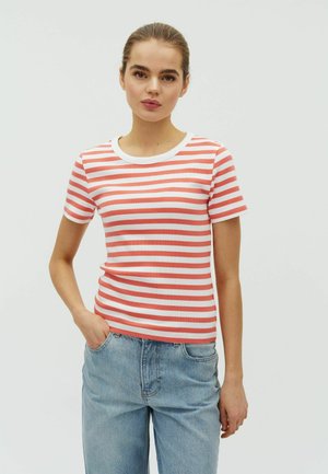 Fiatal nő, hosszú haja hátrakötve, piros-fehér csíkos rövid ujjú ingben és világoskék farmerben, egyik kezét zsebébe dugva áll.