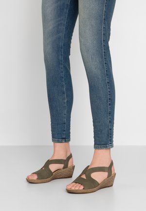 Oliivinvihreät viistosti leikatut sandaalit, joissa on leikkauksia, kuvioitu kudottu korko ja avoin varvas, yhdistettynä kapeisiin sinisiin farkkuihin.