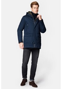 Granatowa pikowana parka z odpinanym kapturem, zapięciem na zamek błyskawiczny z przodu, wieloma kieszeniami i kontrastowym podszyciem. Noszona z ciemnymi dżinsami i brązowymi butami.