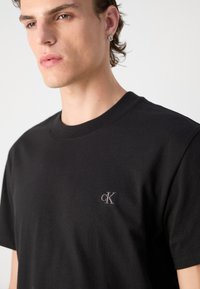 T-shirt nera in cotone con scollo rotondo, caratterizzata da un piccolo logo grigio "CK" sul lato sinistro del petto. Maniche corte e texture morbida.