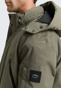 Olivno zelena zimska jakna z visokim ovratnikom, podloženo zasnovo, zadrgo na žepu in črnimi kovinskimi dodatki. Podložena z temnim materialom.