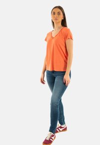 T-shirt orange avec un col en V et des manches courtes, associé à un jean skinny bleu clair et des baskets bordeaux avec des accents blancs. Design simple.