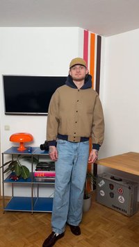 Νέος άνδρας που φοράει καφέ μπουφάν, μπλε τζιν και καπέλο, στέκεται σε ένα δωμάτιο με τηλεόραση, πορτοκαλί λάμπα, φυτά και μια διακοσμημένη ασημένια θήκη.