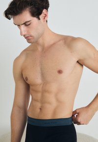 Schwarze, enganliegende Unterwäsche mit einem grauen Calvin Klein Bund, getragen von einem männlichen Modell mit einem durchtrainierten Körperbau, das vor einem neutralen Hintergrund steht.