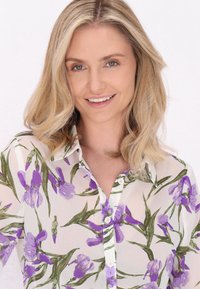 Bluză transparentă cu imprimeu floral, prezentând irisuri violete și frunze verzi. Design cu nasturi, fundal deschis și textură subtilă.
